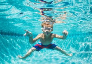 a boy smiling for the camera wearing goggles and a swim suit as he swims underwater in a pool