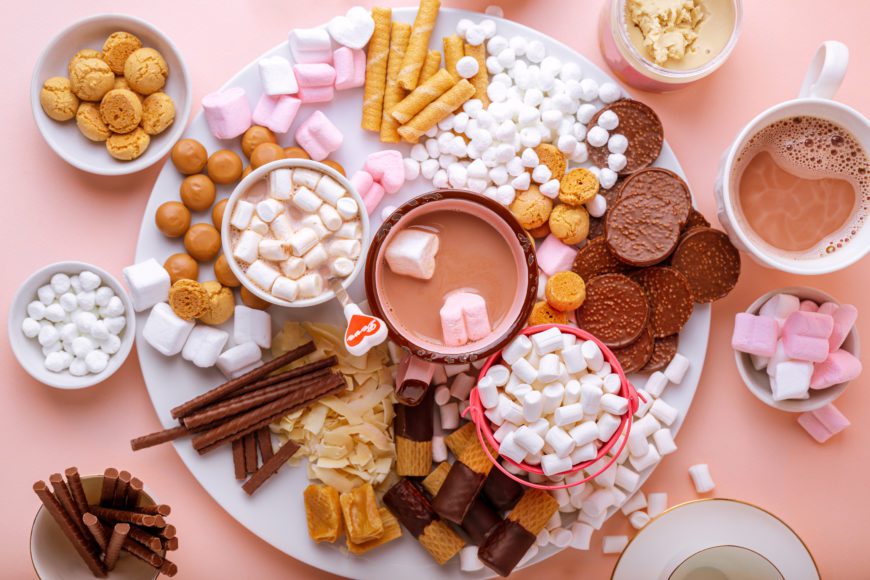 Hot chocolate, marshmallows, chocolates and cookies charcuterie board on pink background. Closeup view, horizontal orientation
