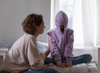 a teenager sitting cross legged on her bed with her hood on, the strings pulled tight to close it as her mom tries to talk to her