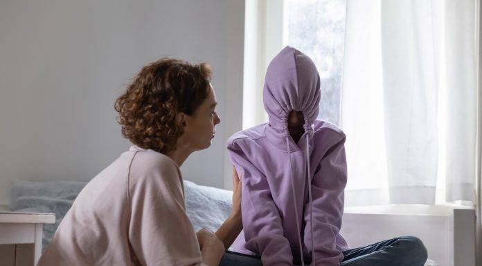 a teenager sitting cross legged on her bed with her hood on, the strings pulled tight to close it as her mom tries to talk to her