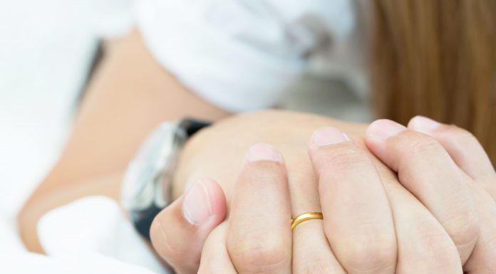 a close up of two people holding hands, one is wearing a wedding ring, as they lay in bed together