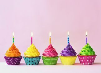 five birthday cupcakes with colorful wrappers and bright colored icing with lit candles on top against a pink background