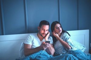 a man and a woman, both sitting up in bed as they share earbuds and listen to a podcast