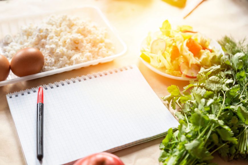an empty notebook and pen on the counter surrounded by healthy foods for meal planning