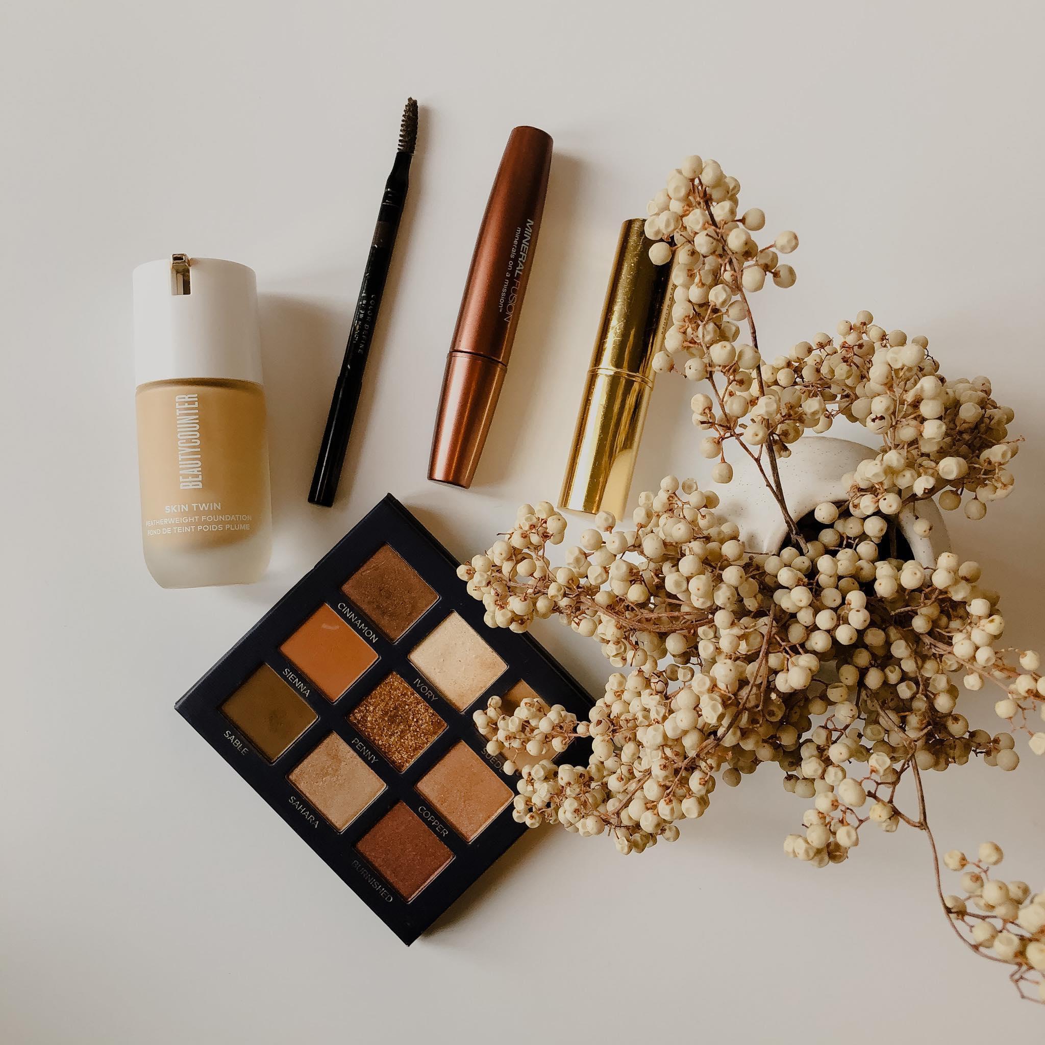 clean skincare and beauty products such as a pallet of brown eyeshadow, mascara, and foundation, on a cream colored background with flowers 