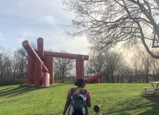 a mom with a baby in a carrier on her back, and her toddler walking next to her as they explore a park