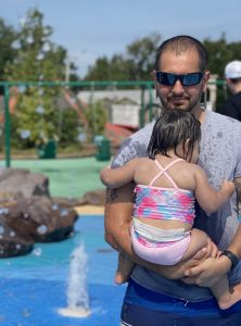a dad in sunglasses holding his little girl in her swimsuit at a playground splash pad