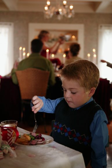 a boy sitting at the kids’ table at the holidays, with the adult table in the background