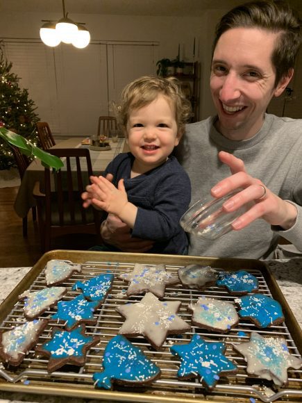 a dad holding a toddler on his lap as they decorate Hanukkah sugar cookies