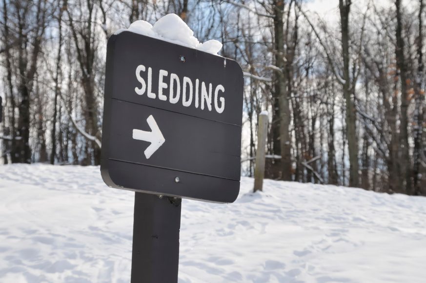 a sign with an arrow that says “sledding” on a snowy hill near the woods
