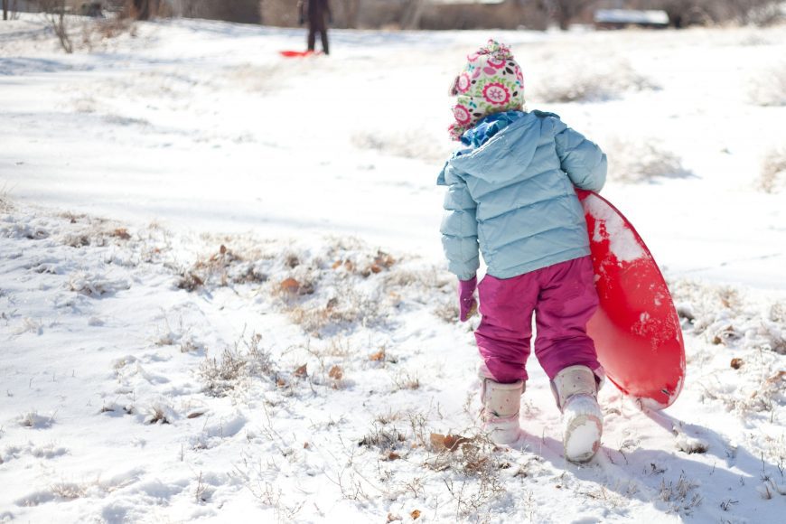 a young girl in a light blue jacket and pink snow pants walking in the snow with her red plastic sled