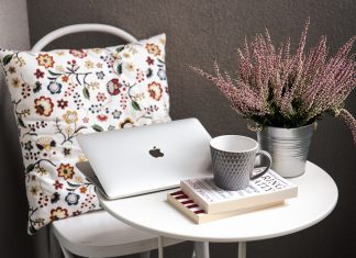 a MacBook on a small white table with two books, a coffee cup, and a plant