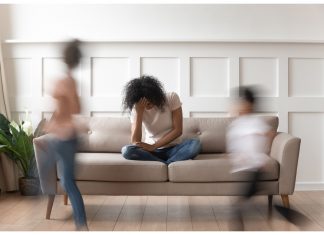 a mom sitting on the couch with her head in her hands as her kids run in circles around her