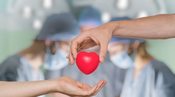 an operating room out of focus in the background with a hand in the foreground, handing a plastic heart to an open palm