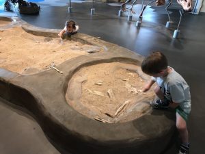 A boy and a girl playing in a sand pit shaped like a dinosaur bone at The Museum at Prairiefire in Kansas City