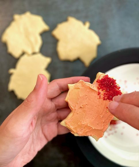 a leaf shaped sugar cookie with orange icing as someone is spreading sprinkles on top of it