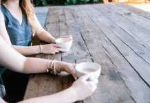 a close up of two women holding coffee cups on a long wooden table