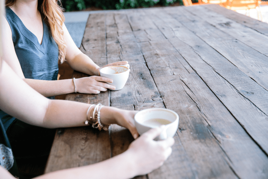 a close up of two women holding coffee cups on a long wooden table