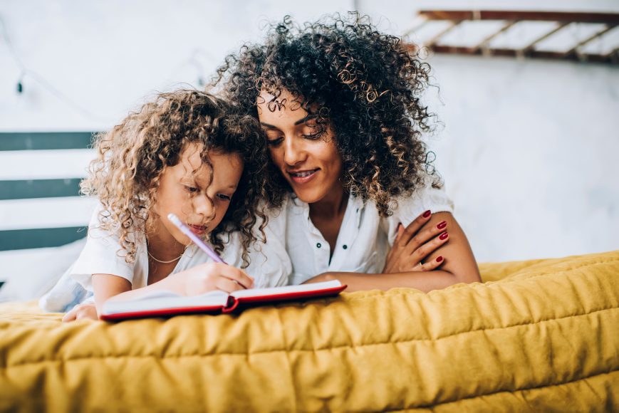 African American Mother and daughter with afro hair lying in bed together and concentrated on writing in journal while smiling