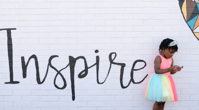 The word “inspire” painted on a white brick wall as a young girl stands next to it