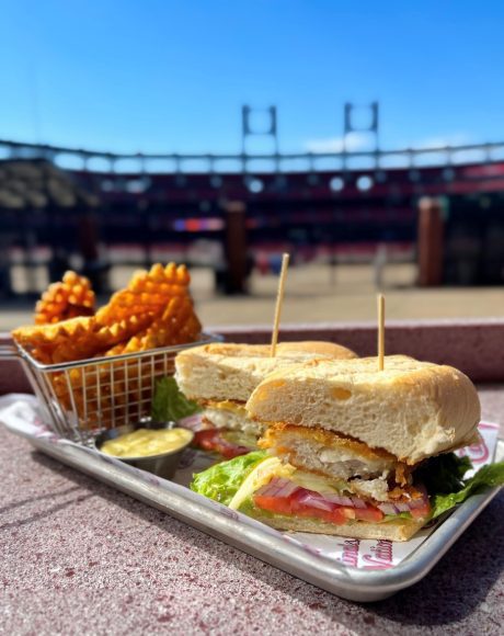 a breaded fish sandwich and sweet potato fries on a platter with Busch Stadium in the background