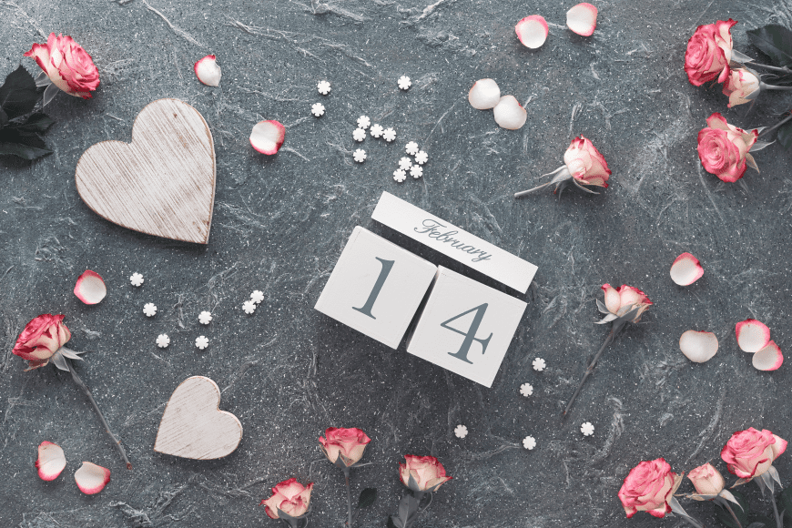 a wooden block calendar on the date of February 14 on a gray surface covered with hearts and pink flowers