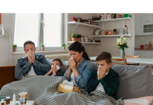a mom, dad, and their two kids all under a blanket on the couch. They all have tissues to indicate they are sick with viruses
